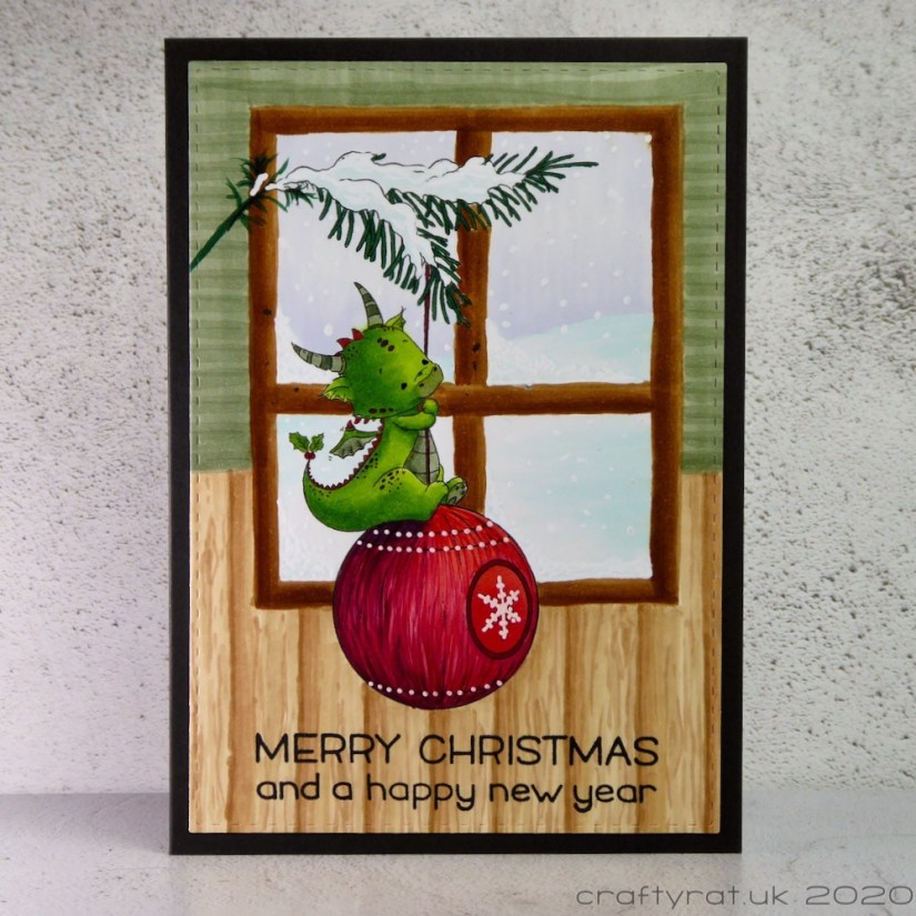 A Christmas card with a little green dragon sitting on a red bauble hanging from a sprig of fir in front of a window.