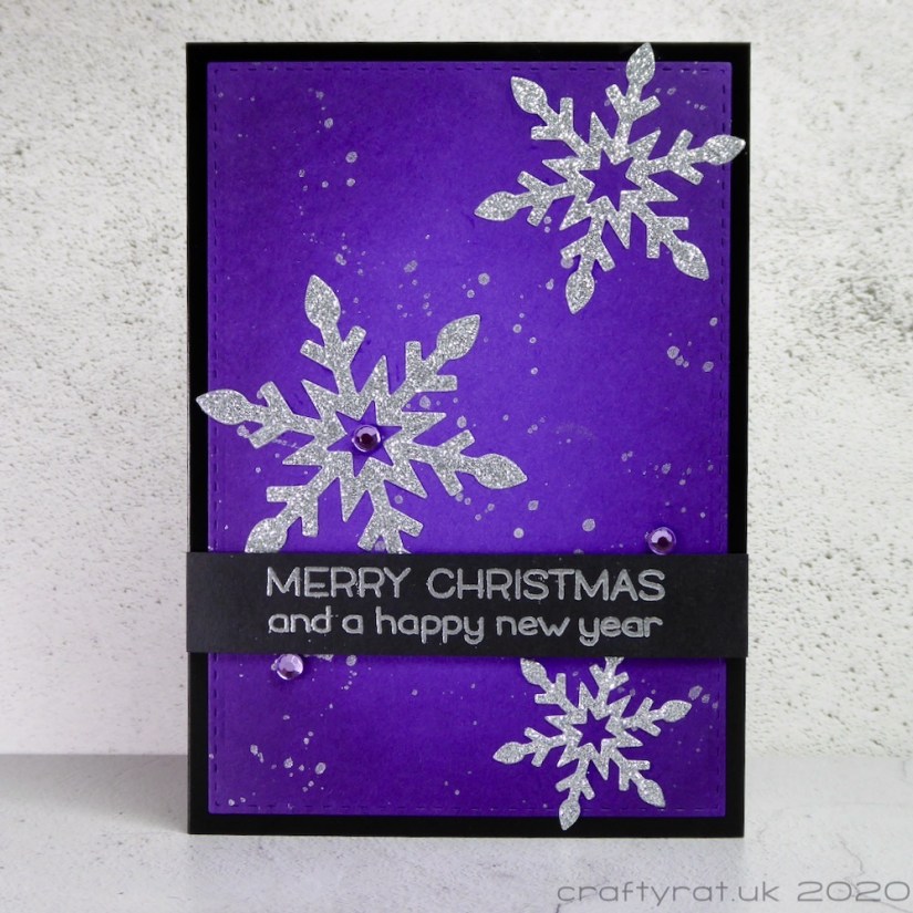 A Christmas card with silver glitter snowflakes on a purple background splattered with silver paint.