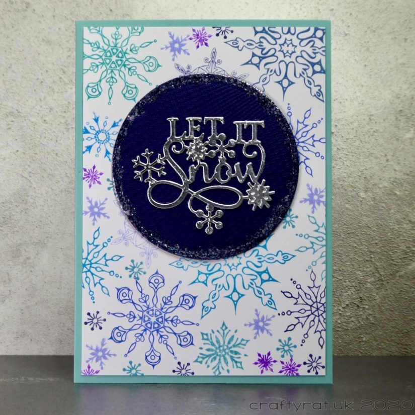 A Christmas card with a background covered in detailed snowflakes stamped in blues and purples. On top of that is a dark blue circle with a decorative silver die-cut that says "let it snow".