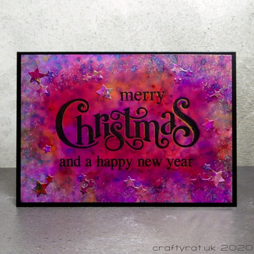 A card that says Merry Christmas and a happy new year on top of a red-purple alcohol ink background with die-cut stars.