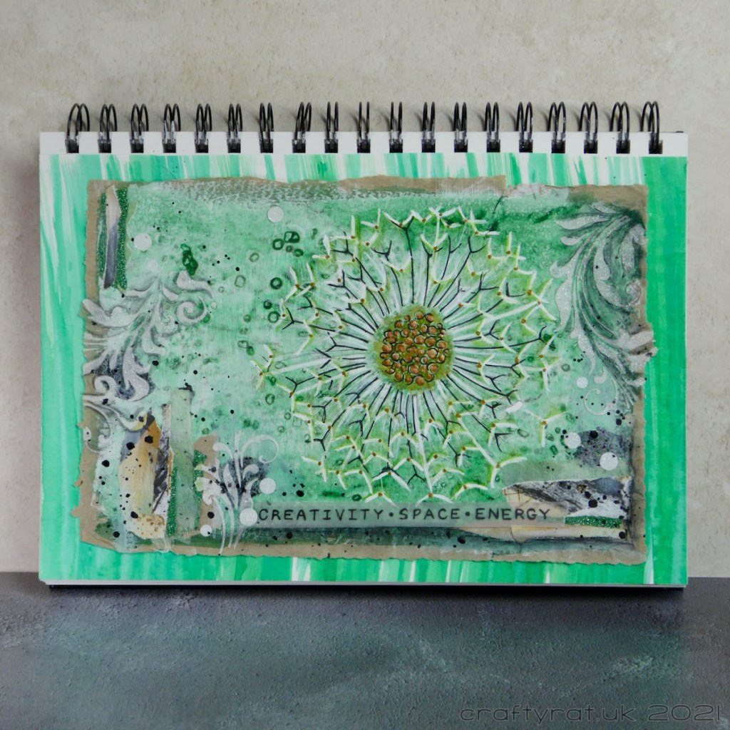 A predominately green art journal page with a dandelion seed head surrounded by collage, stencilled texture paste and a banner reading "creativity - space - energy".